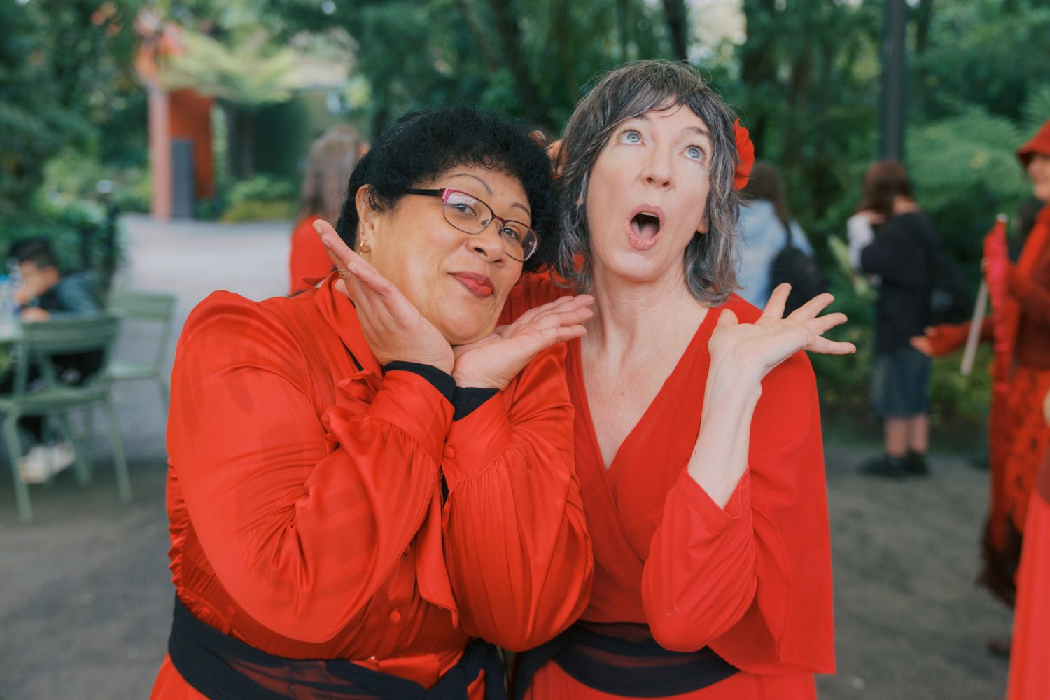 Two women in matching red costumes pose playfully outdoors, making expressive faces at a garden event.