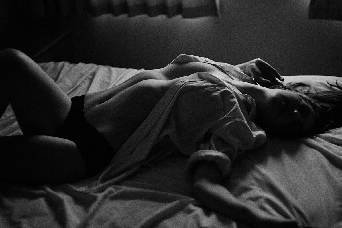 Black and white artistic boudoir photo of a woman reclining on a bed in soft dramatic lighting.