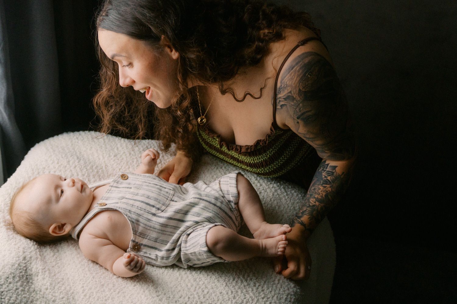 Tattooed mother lovingly gazing at her newborn baby lying on a white blanket in a dark studio setting.