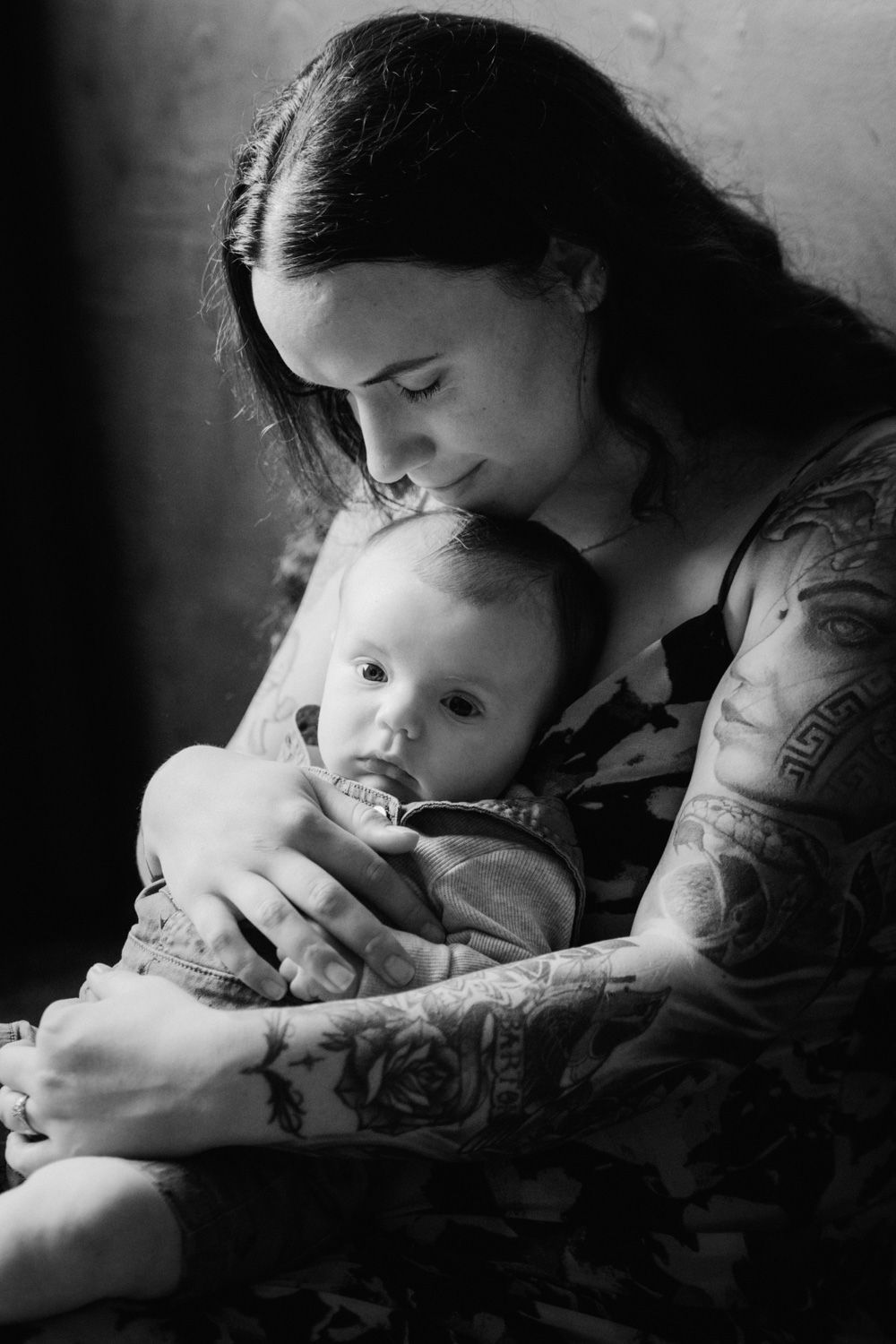 Black and white photo of a tattooed mother tenderly holding her baby, gazing down with love.