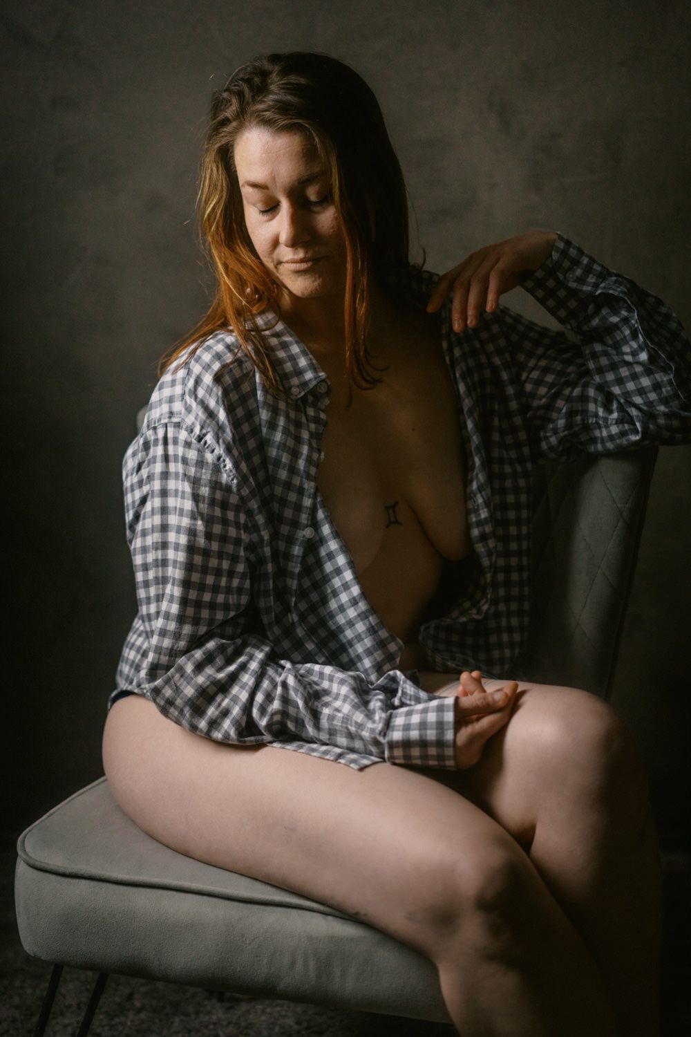 Woman in open plaid shirt seated on chair in moody studio portrait.