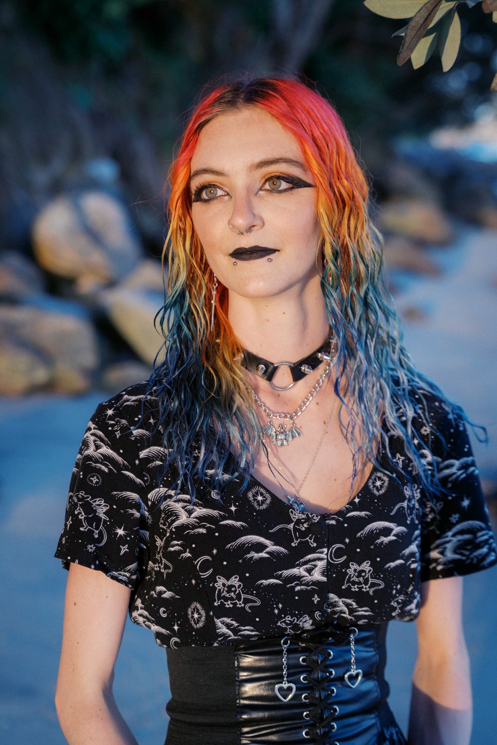 Young woman with rainbow-dyed hair, black lipstick, choker necklace, and celestial print top poses outdoors.