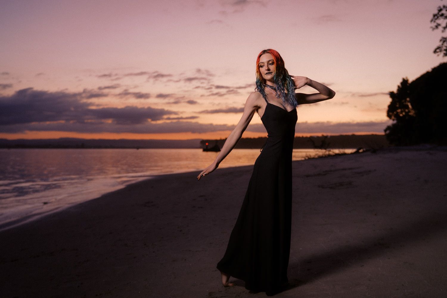Woman in black gown poses on beach at sunset with purple sky and calm water in background.