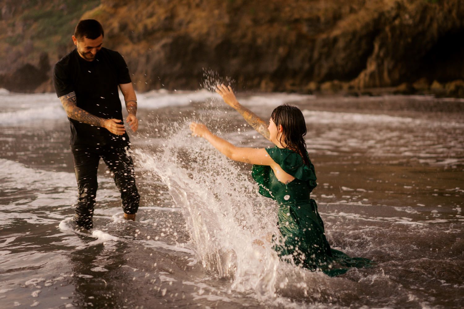 Couple playing in ocean waves on a rocky beach, woman in green dress splashing water at tattooed man in black outfit.