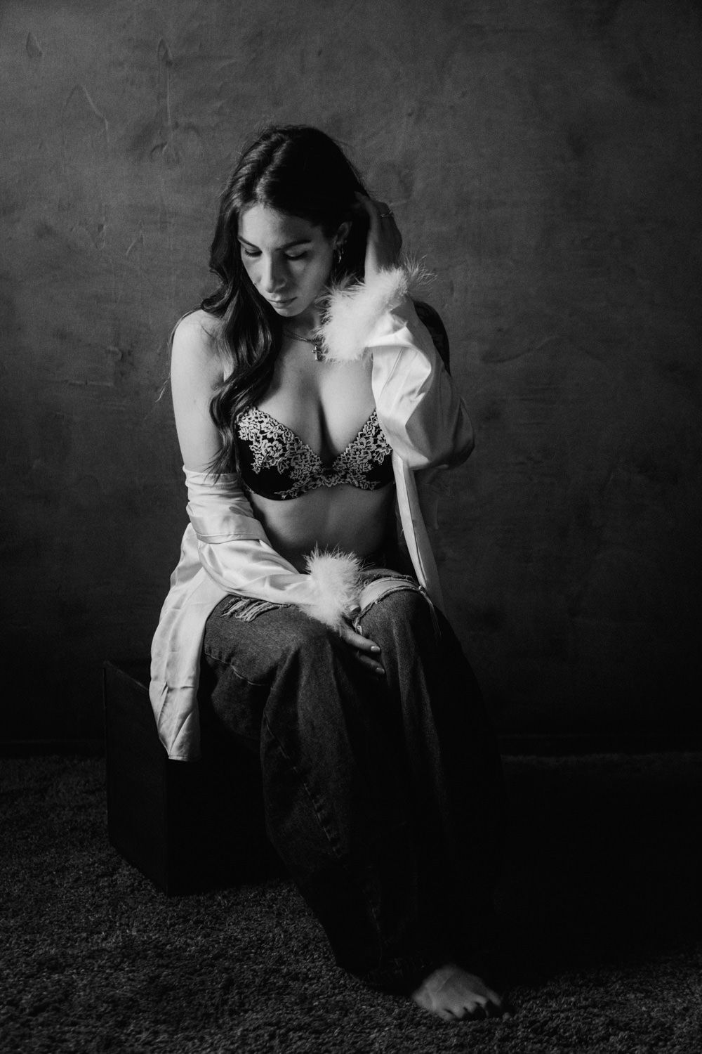 Black and white studio portrait of a woman in lace bra and open robe sitting pensively on the floor.