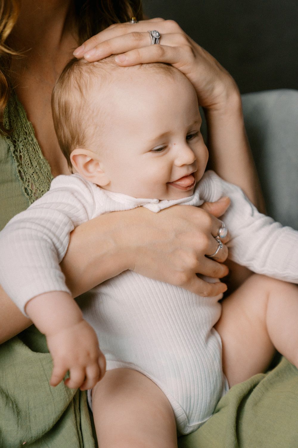 A smiling baby in a white onesie being cradled by a mother wearing a green dress and diamond rings.