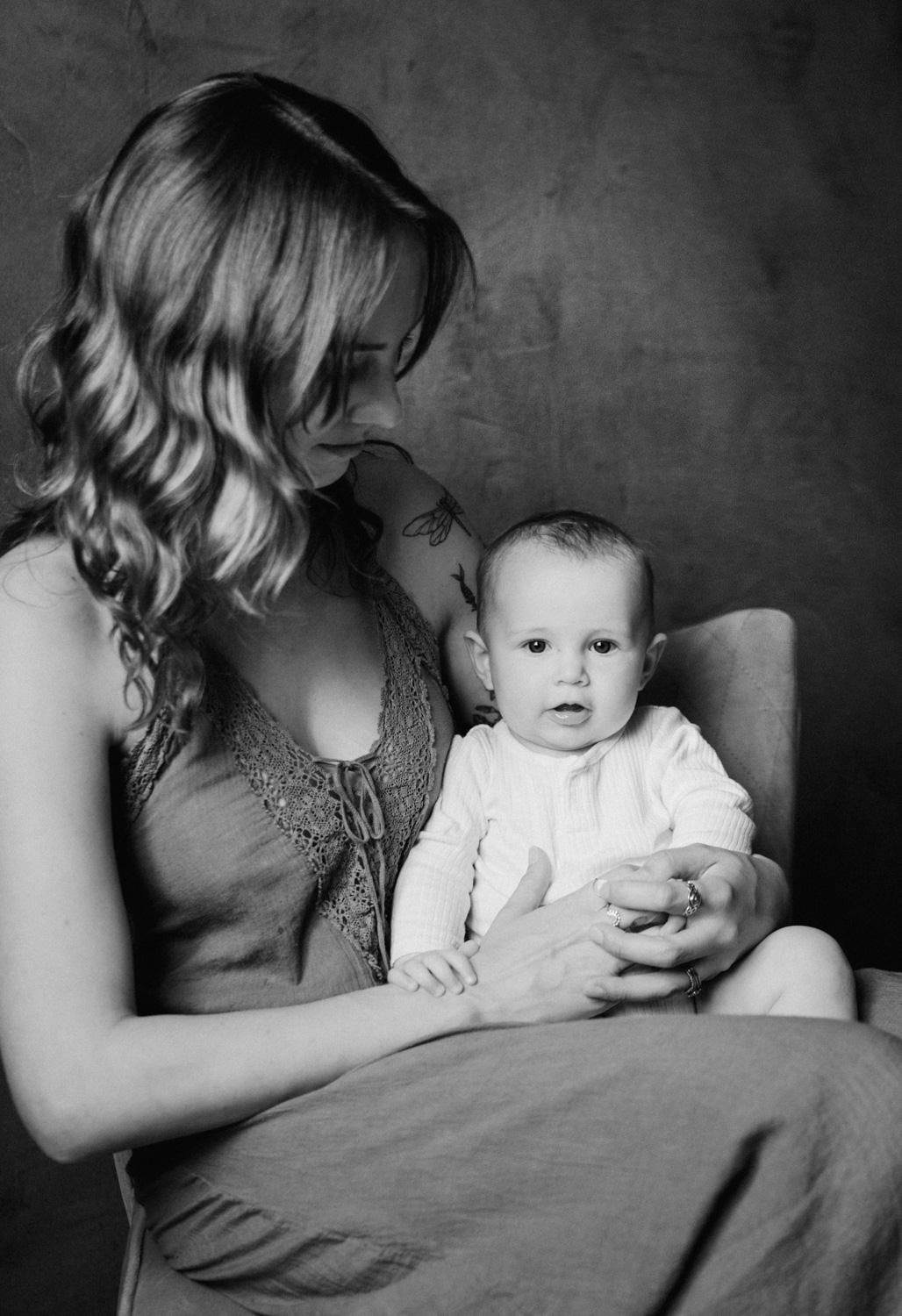 Black and white portrait of a mother holding her baby infant while seated in a chair, looking down lovingly.