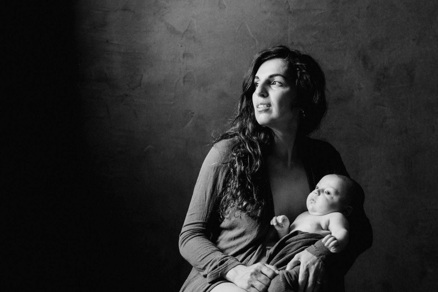 Black and white portrait of a mother holding her newborn baby, gazing thoughtfully in soft studio lighting.