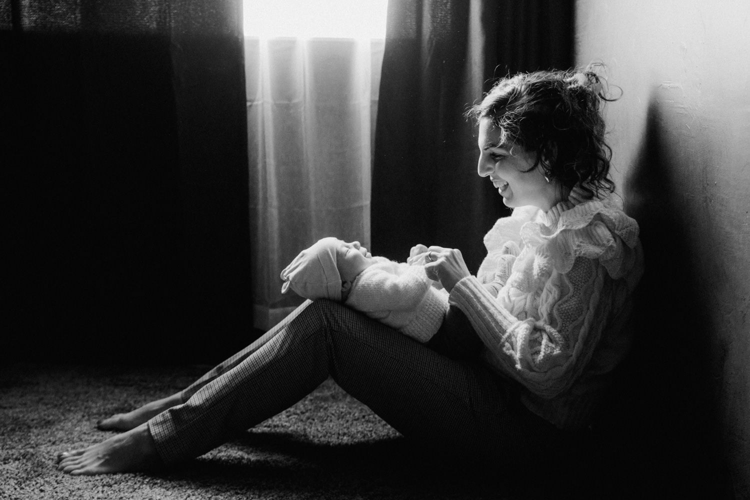 Black and white photo of a young girl sitting alone on the floor, hugging her knees near a window.