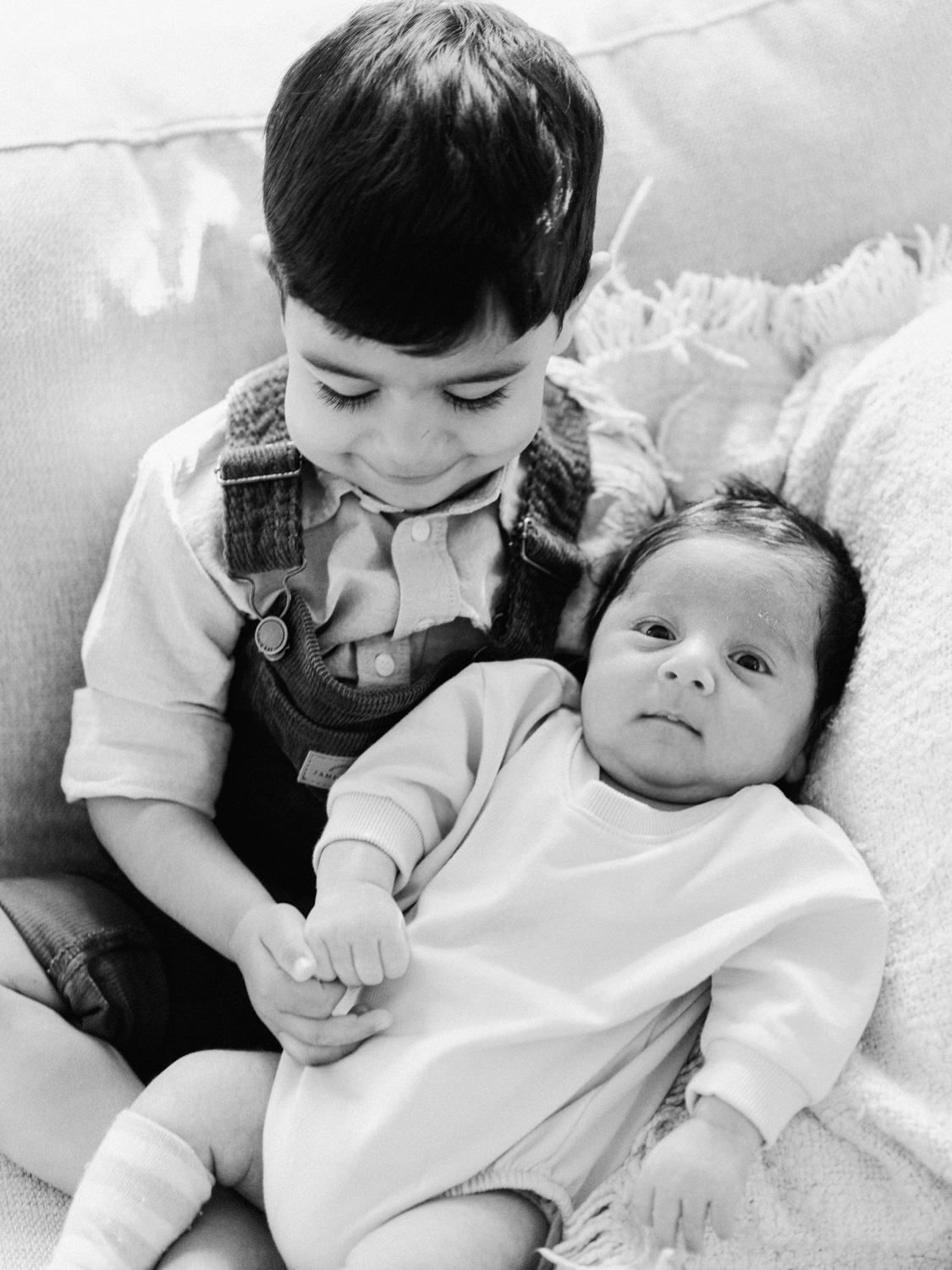 A young boy smiles while holding his newborn sibling in a black and white portrait photo.