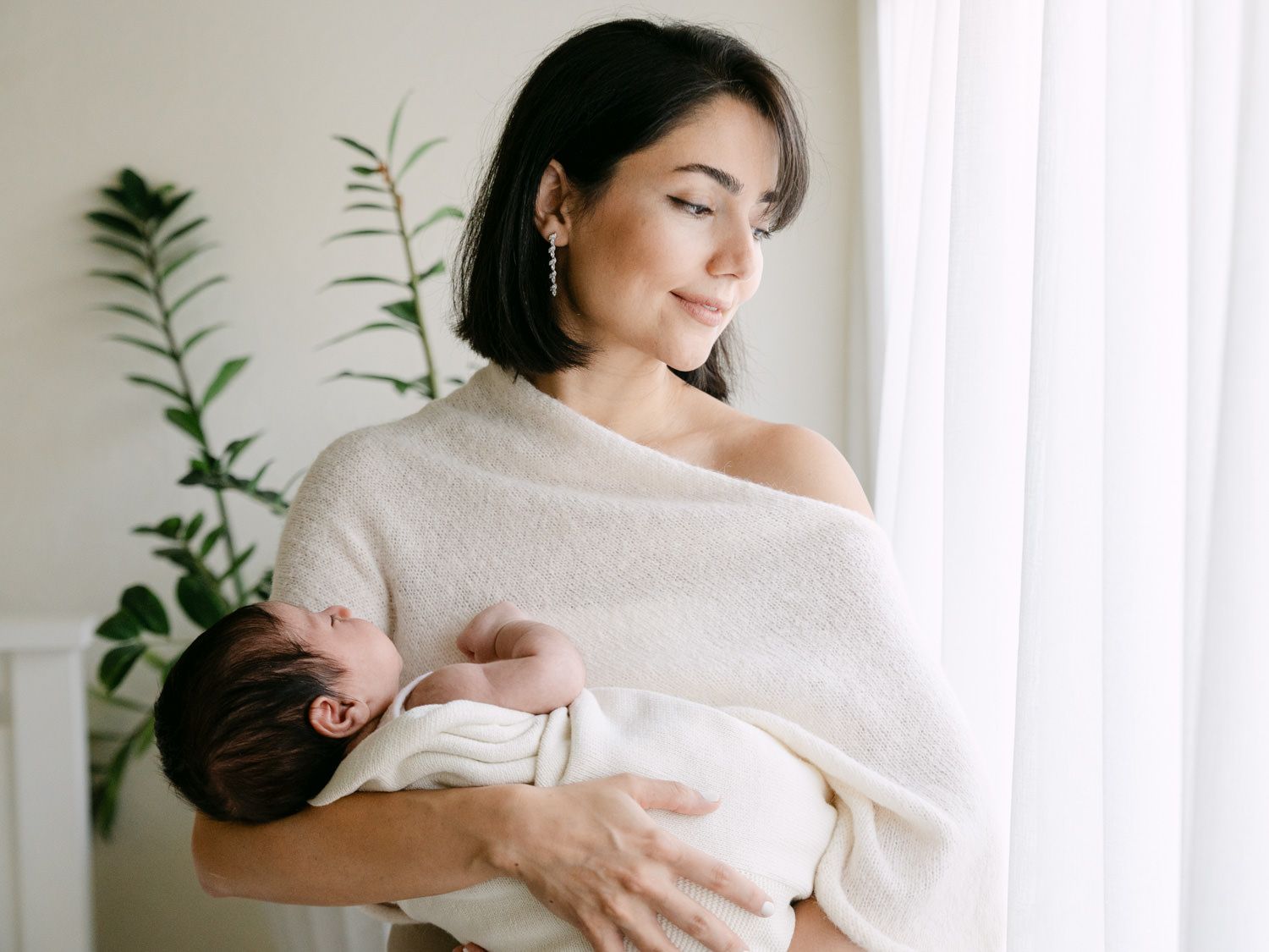 Dark-haired mother holding her swaddled newborn baby near a sunlit window.