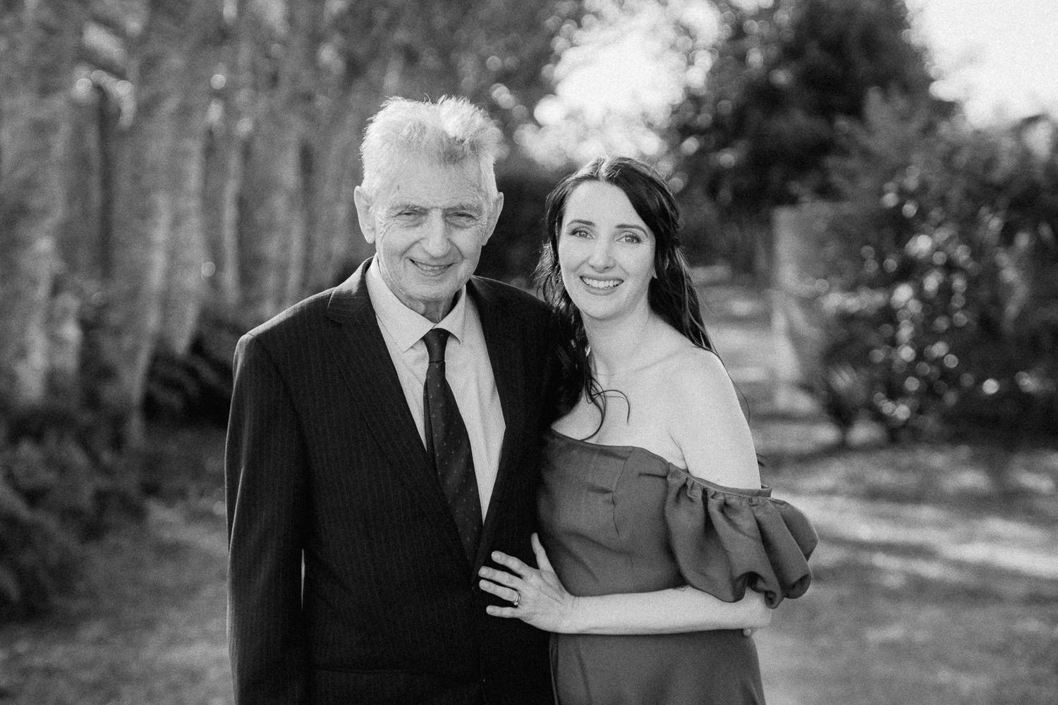 Black and white portrait of an older man and young woman smiling together outdoors among trees.