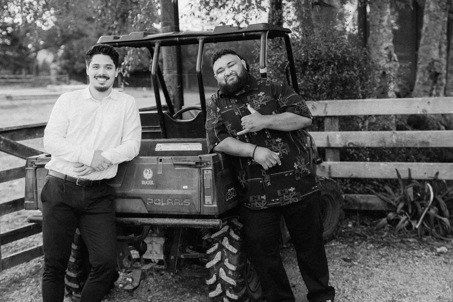 Two men pose on a utility vehicle in a park setting in this black and white photograph.