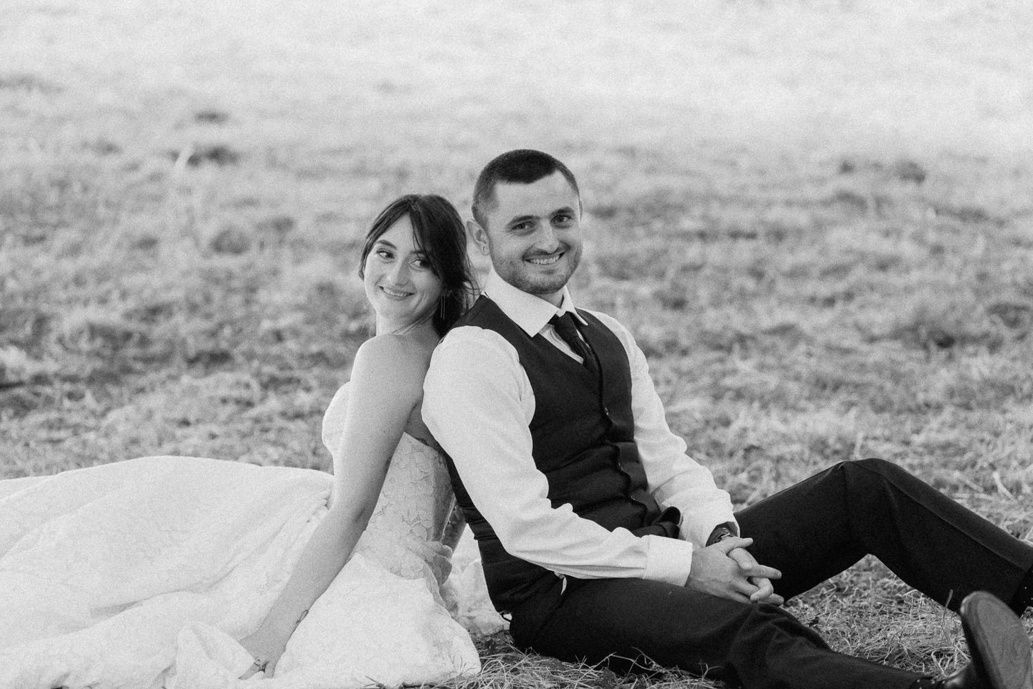 Black and white photo of a bride and groom sitting back-to-back on grass, smiling on their wedding day.