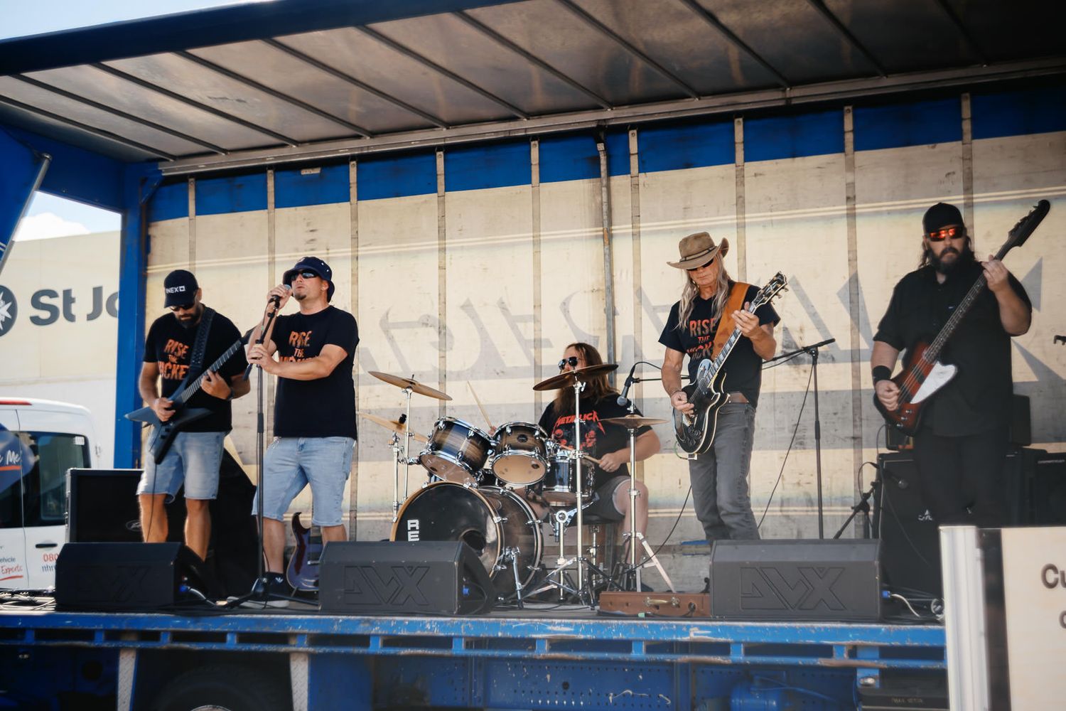 Rock band performing on outdoor stage with guitars and drums at a St. John event on a sunny day.