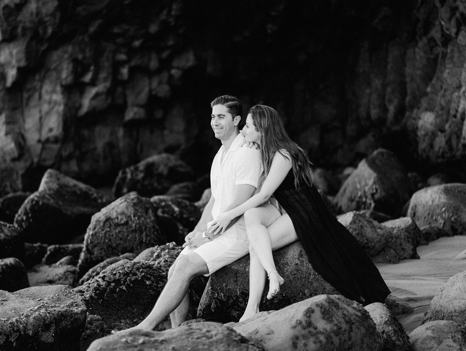 Black and white photo of a romantic couple sitting on coastal rocks near a cliff, embracing during a beach engagement session.