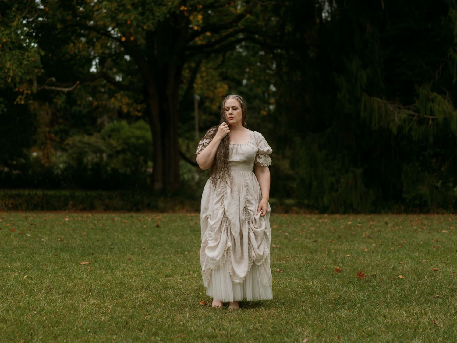 Woman in elegant vintage white gown standing on green grass surrounded by lush trees in an outdoor park setting.