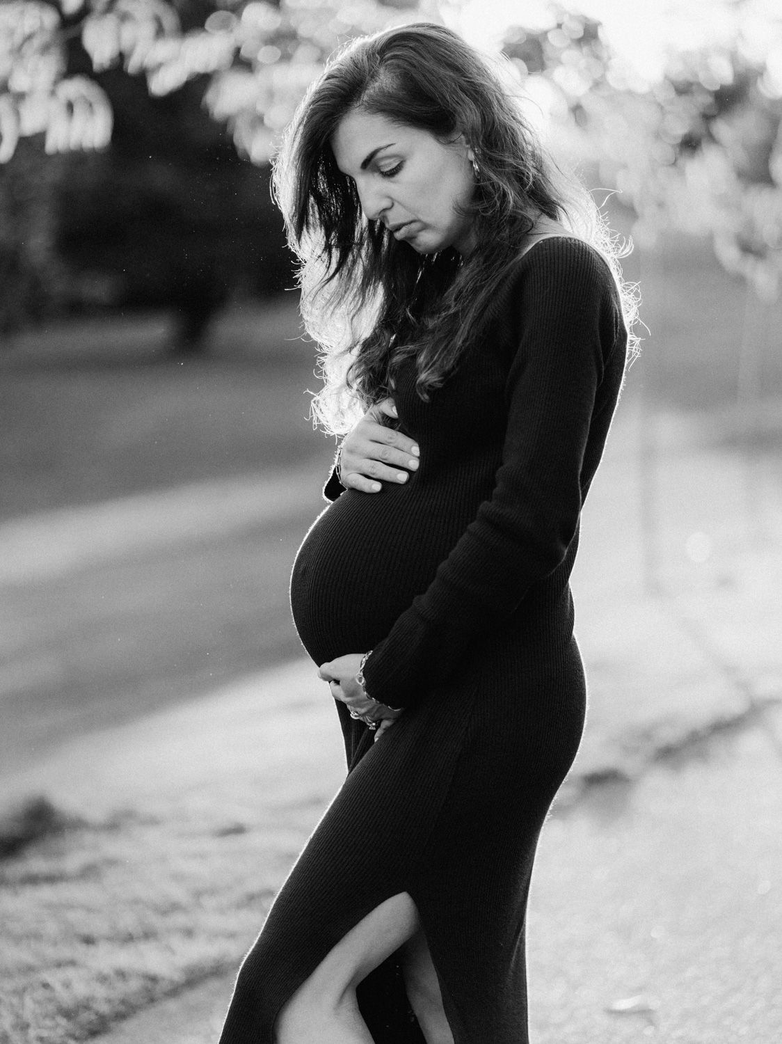Black and white photo of a pregnant woman in a black dress cradling her baby bump outdoors at sunset.