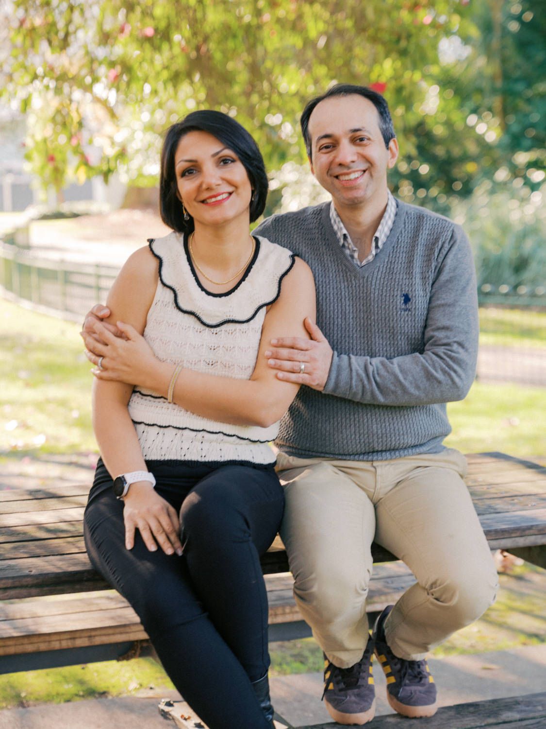 A smiling couple sits together on a wooden bench in a park, the woman in a white top and the man in a gray sweater.