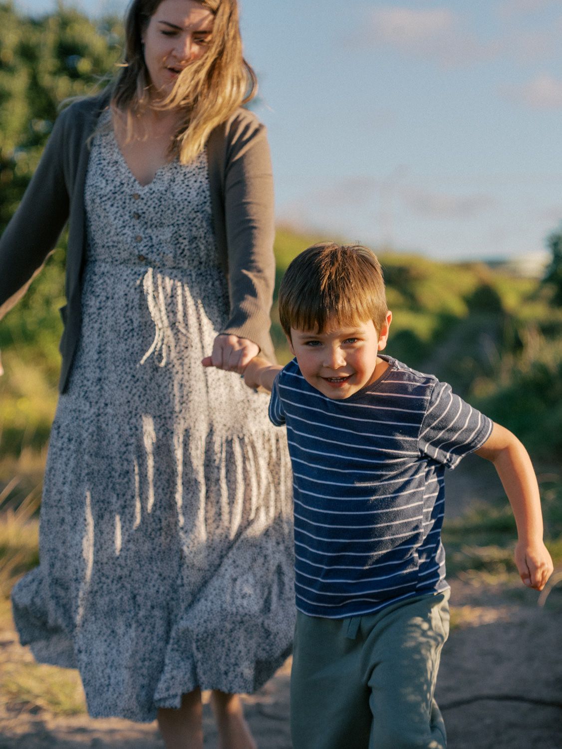 A young boy holds his mother's hand while running along a sandy path surrounded by greenery.