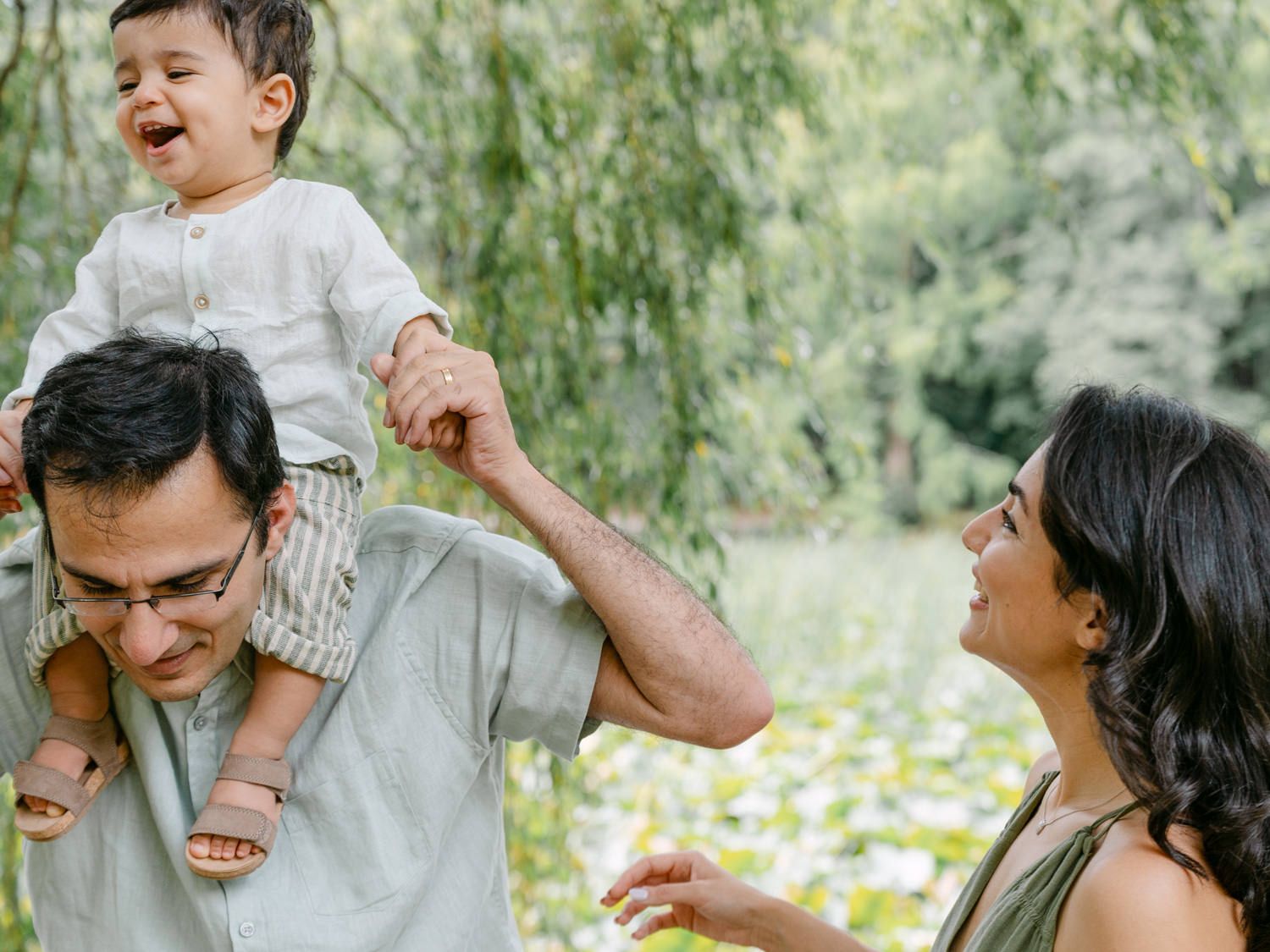 A joyful family outdoors, father carrying laughing toddler on shoulders while mother smiles and holds child's hand.