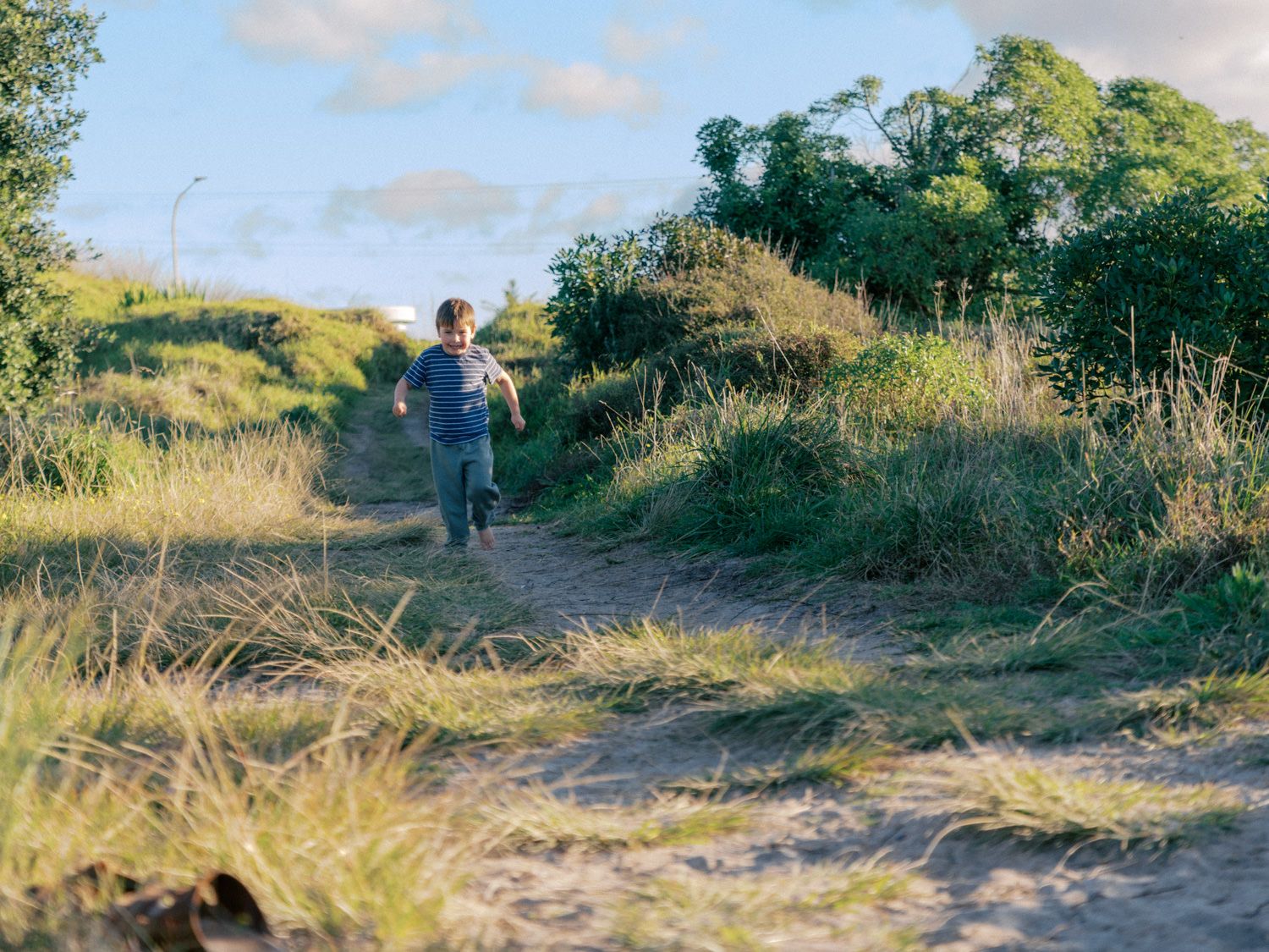 A young child runs along a sandy nature trail surrounded by lush green grass and trees on a sunny day.