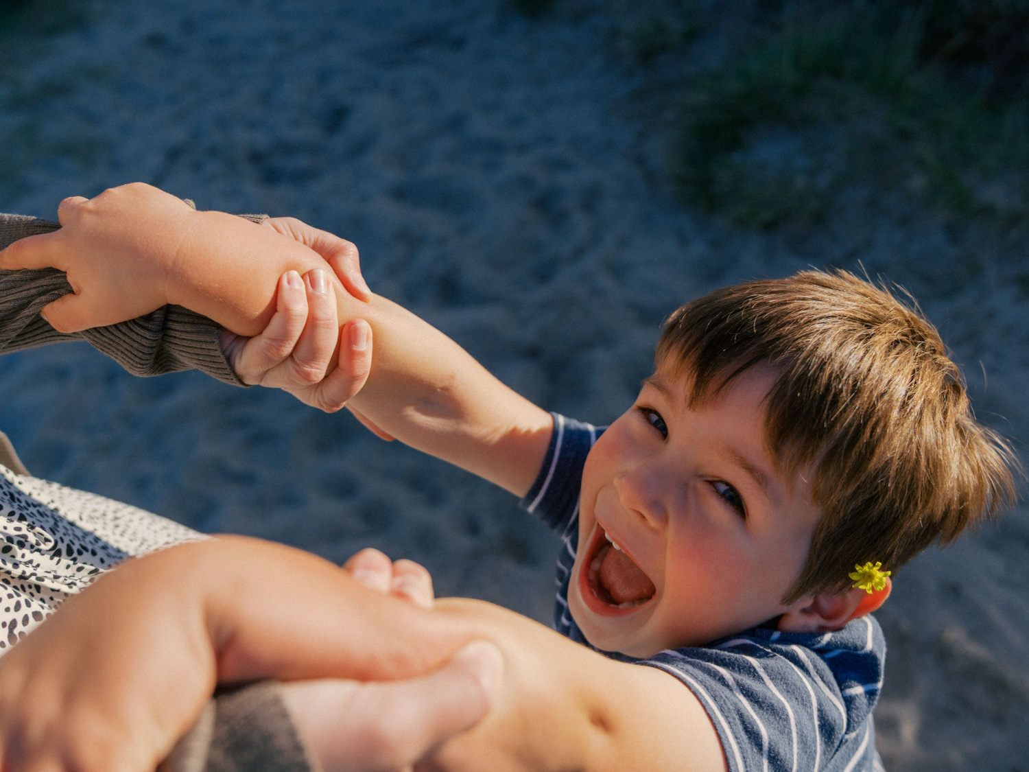A joyful young boy laughs while holding an adult's hands at a sunny beach, wearing a blue striped shirt.