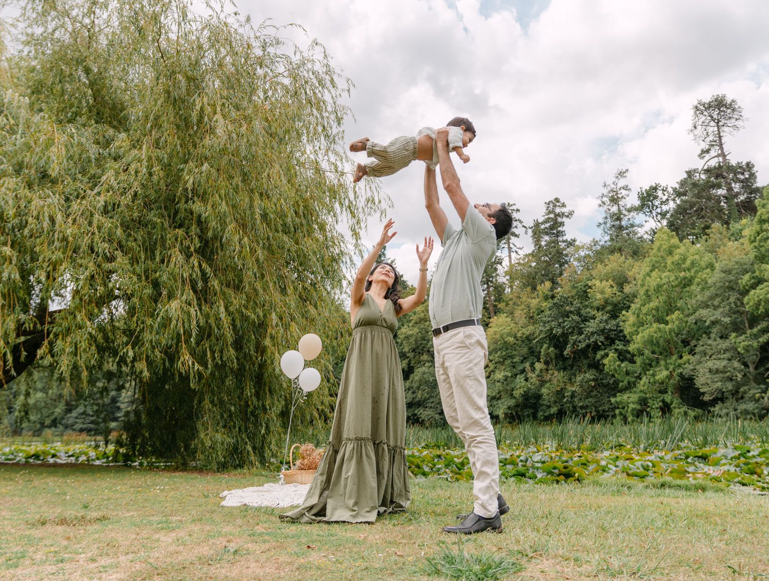 A joyful family outdoors, father tossing baby in the air while mother watches, surrounded by lush green trees and lawn.