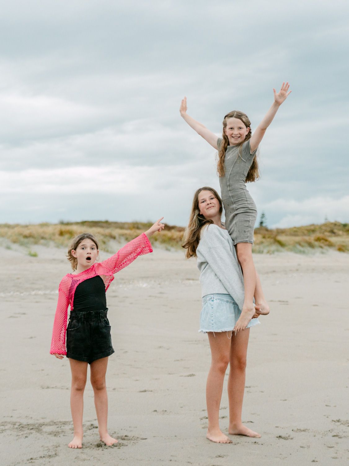 Three girls playing on a sandy beach, one lifted on shoulders with arms raised, under a cloudy sky.