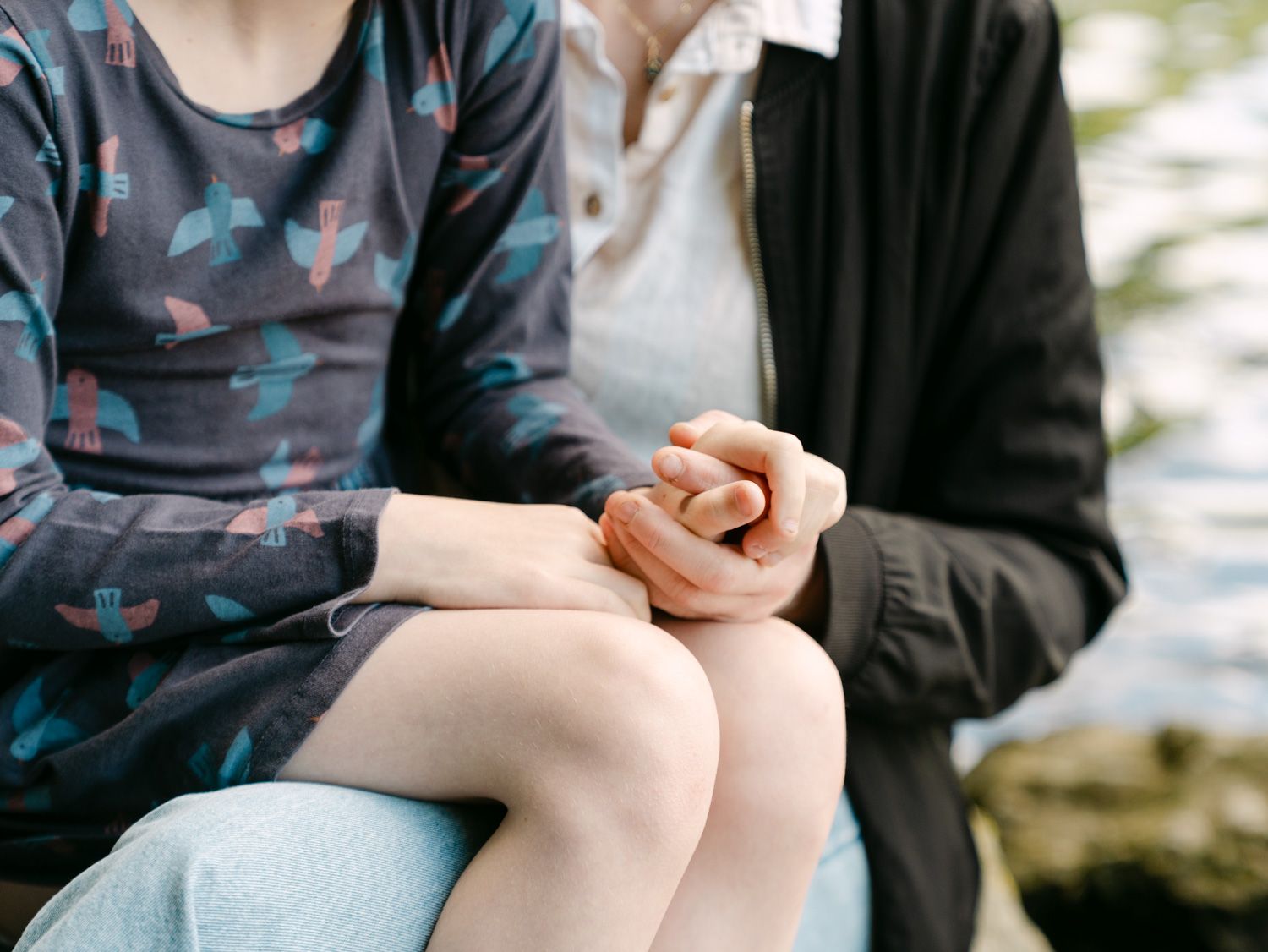 Two people sitting closely together holding hands outdoors, wearing casual clothing near water.