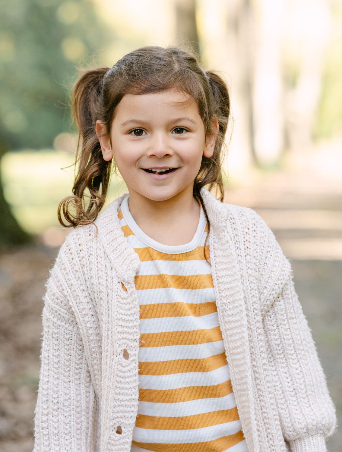 Smiling young girl with pigtails wearing a yellow striped shirt and cream knit cardigan outdoors.