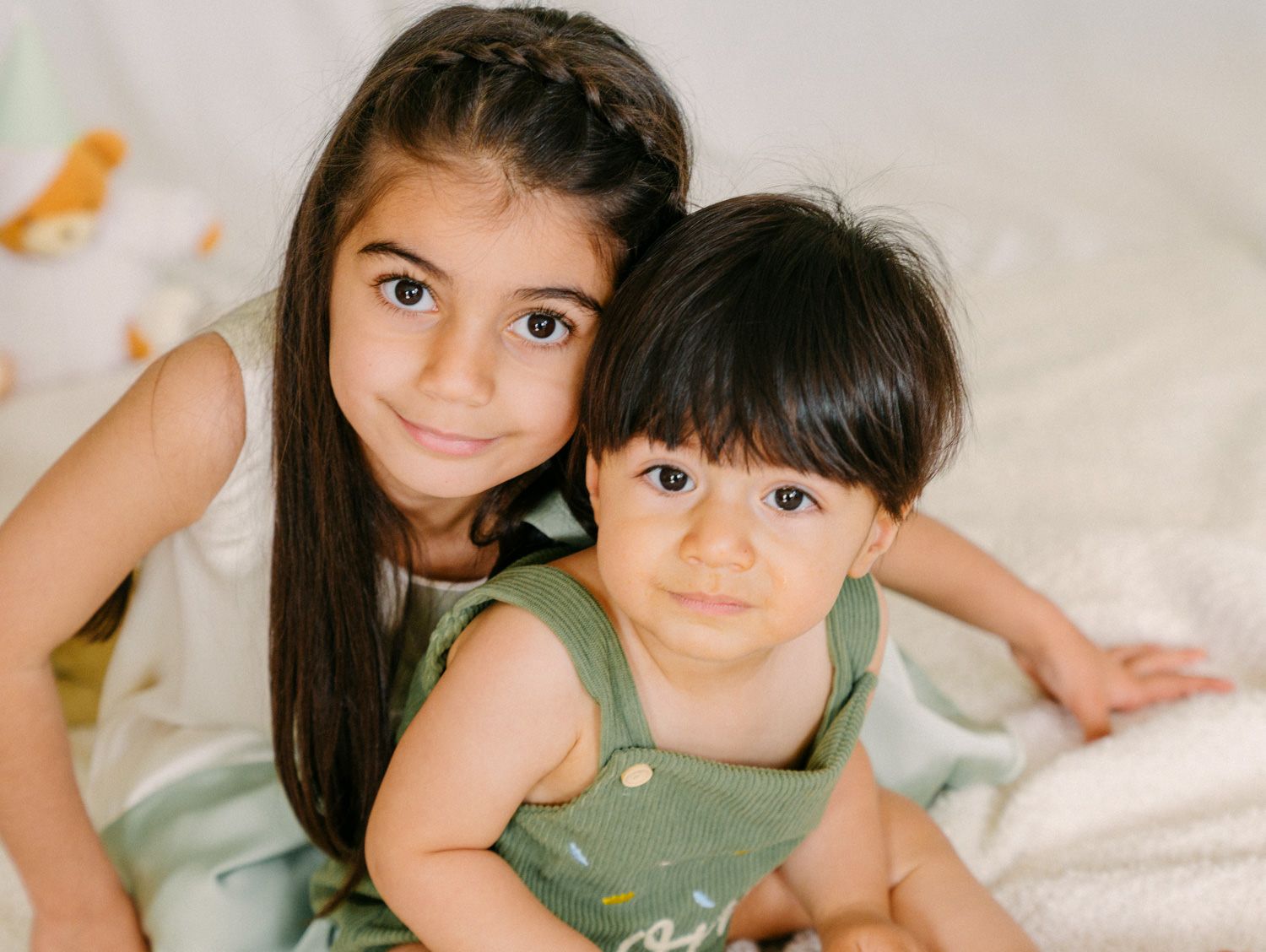 Two young siblings, an older girl with long dark hair and a baby in green overalls, pose together for a portrait photo.