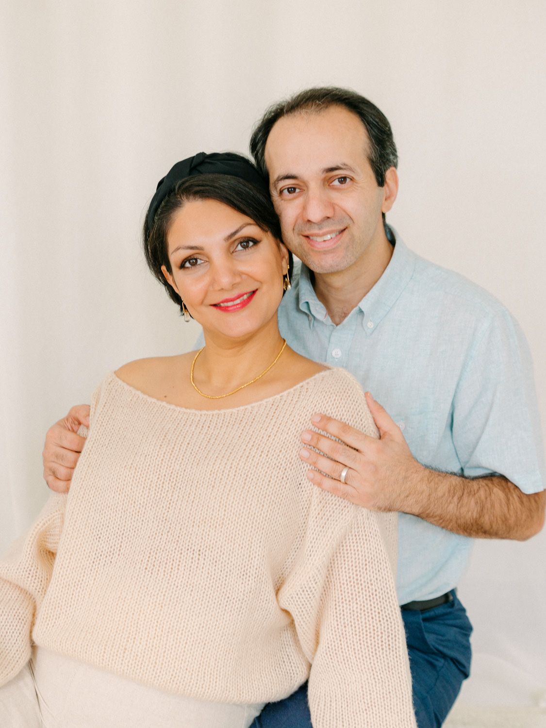 A smiling couple poses together, woman in cream off-shoulder sweater and man in light blue shirt, against white background.