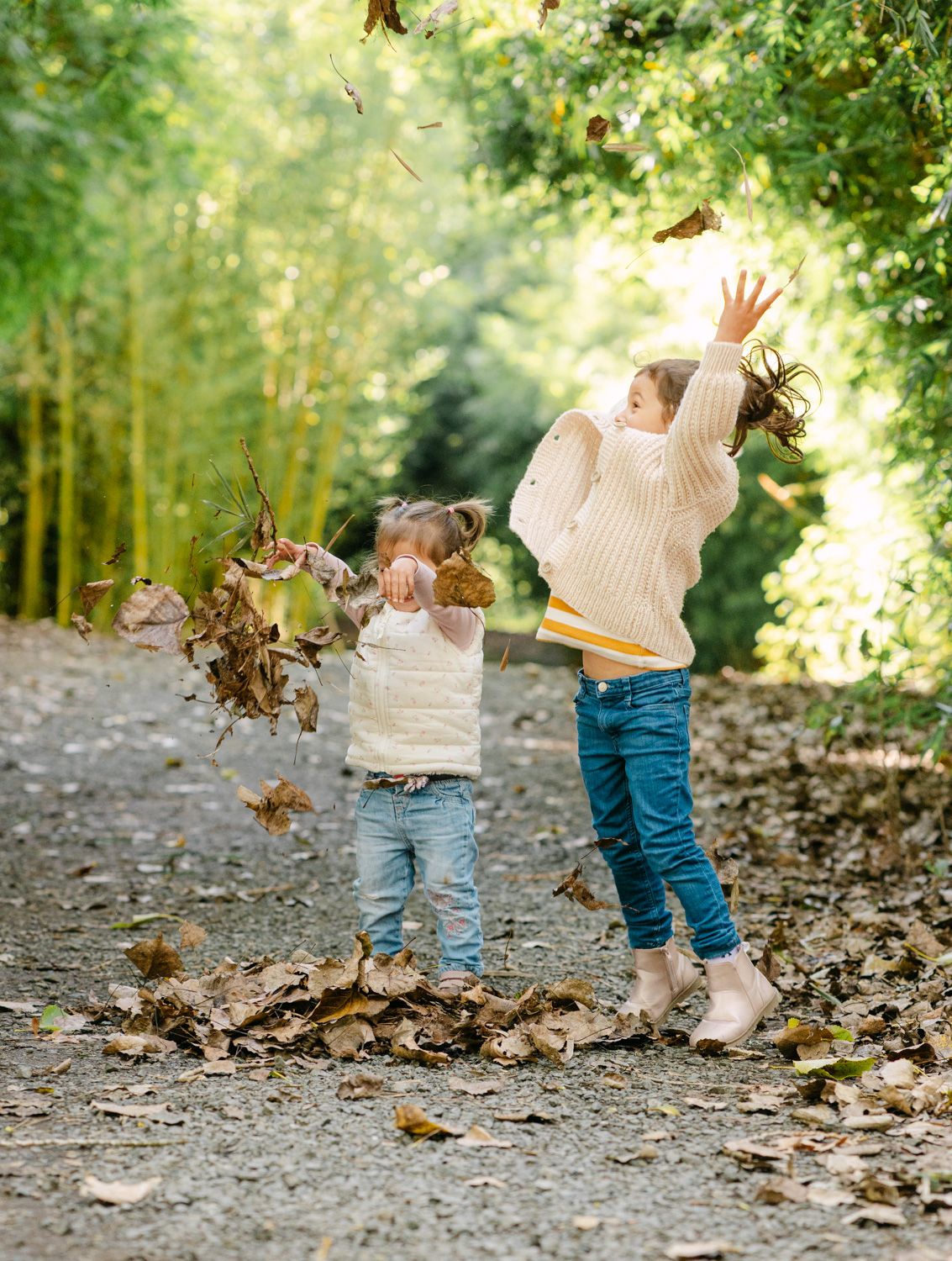 Two young children joyfully tossing autumn leaves in a sunlit forest path, wearing cozy sweaters and jeans.