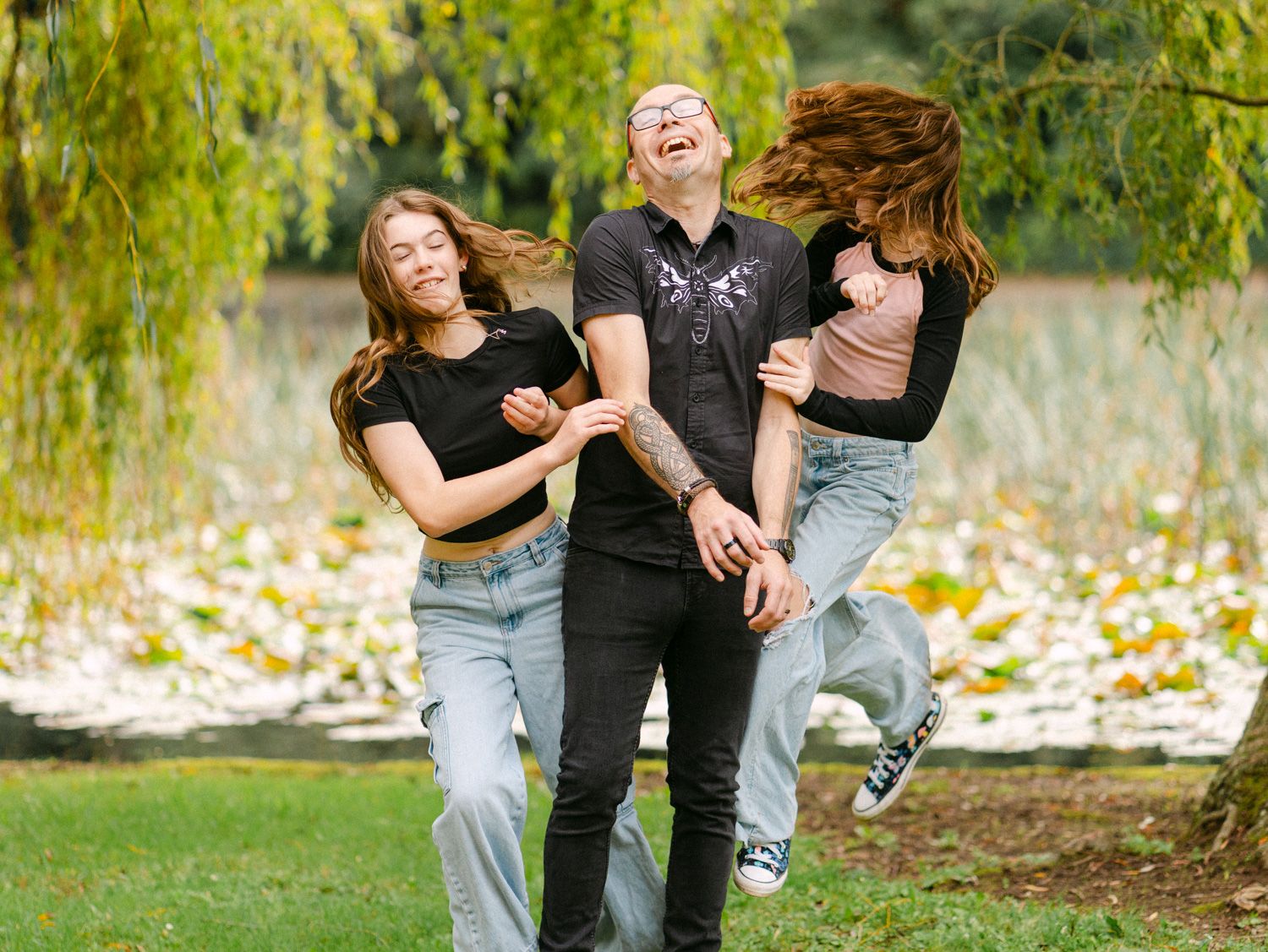 Three people laughing and playing in a park under a weeping willow tree on a sunny day.
