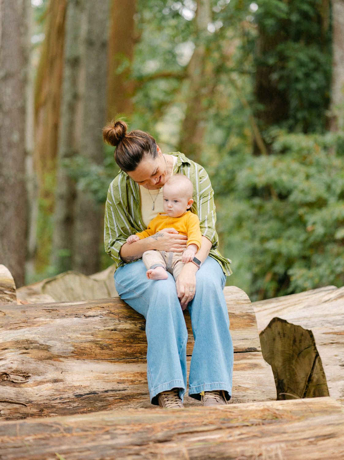 Mother sitting on log cradling baby in yellow outfit in forest setting.