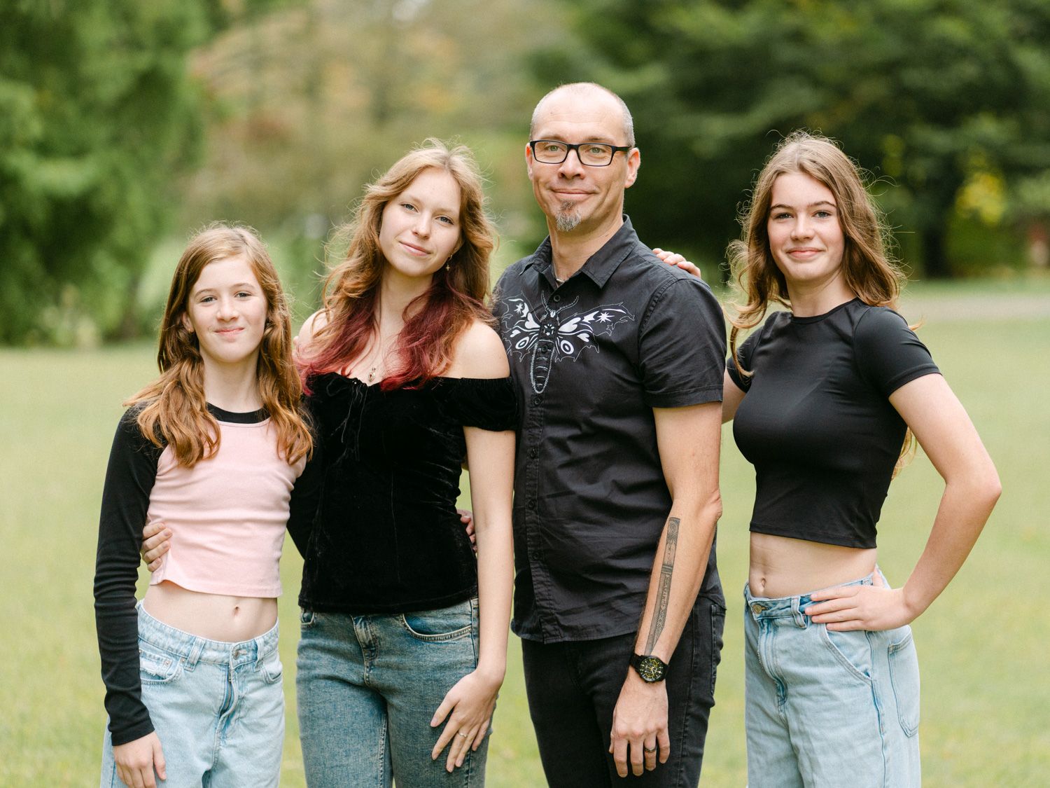 A bald man poses outdoors with three teenage girls in casual attire on a green lawn with trees in background.