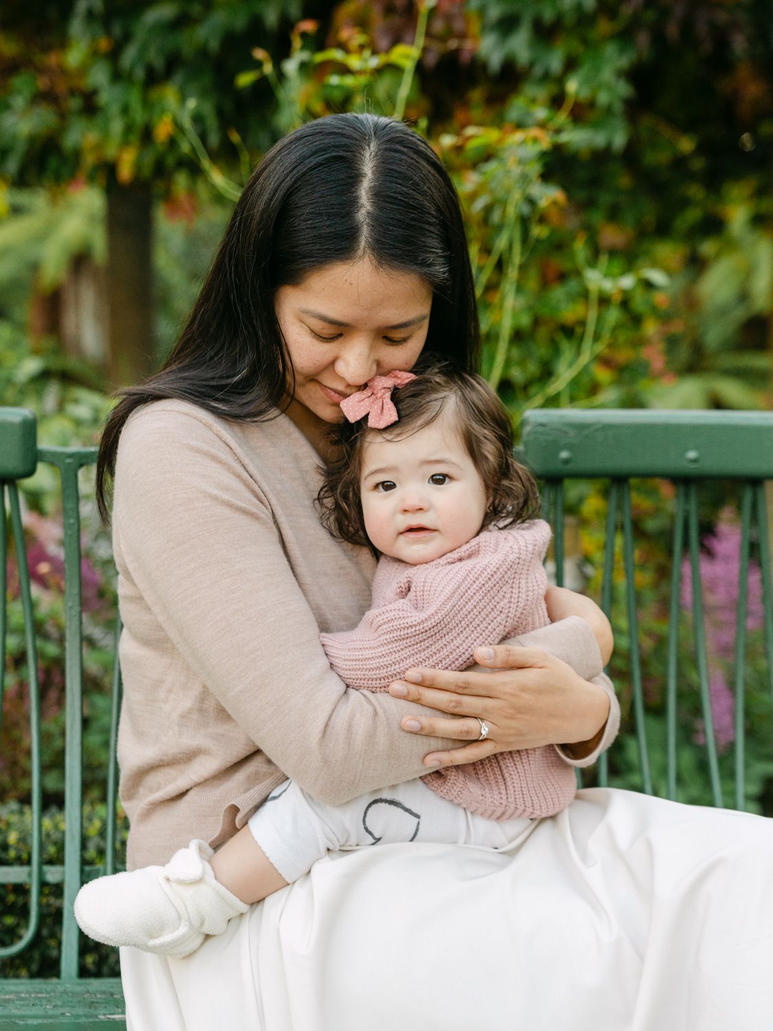 A mother tenderly hugs her baby girl in a lush garden, both wearing soft pink outfits in a warm family portrait.