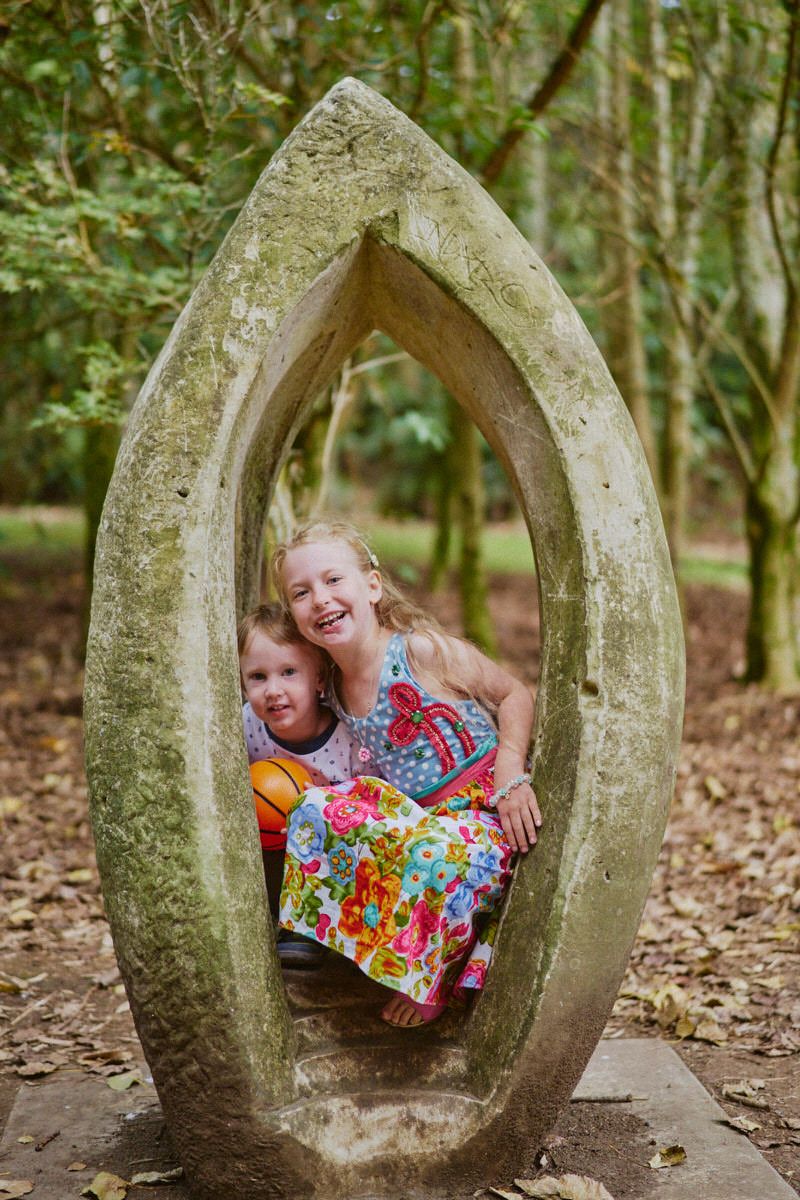 Two children smiling inside a stone arch sculpture in a woodland garden setting.