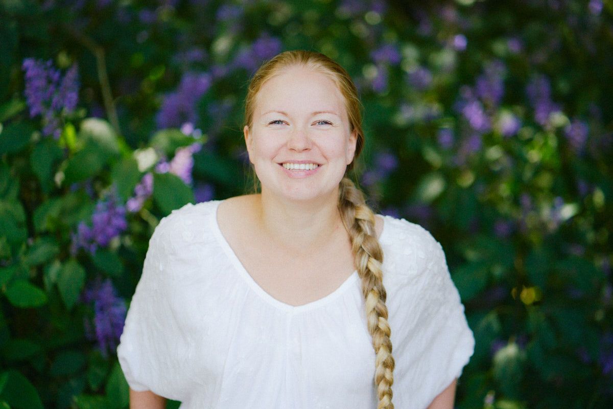 Smiling young woman with a blonde braid wearing a white top, posing outdoors in front of purple lilac bushes.