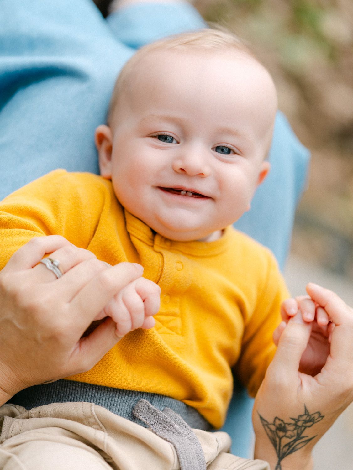 Smiling baby in yellow shirt being held by adult with tattoo and ring outdoors.