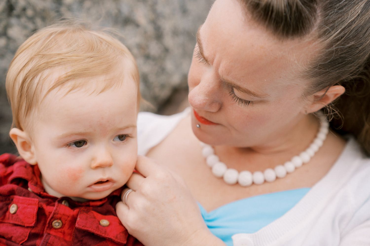A mother in a pearl necklace and blue top tenderly holds her blonde toddler dressed in a red shirt.