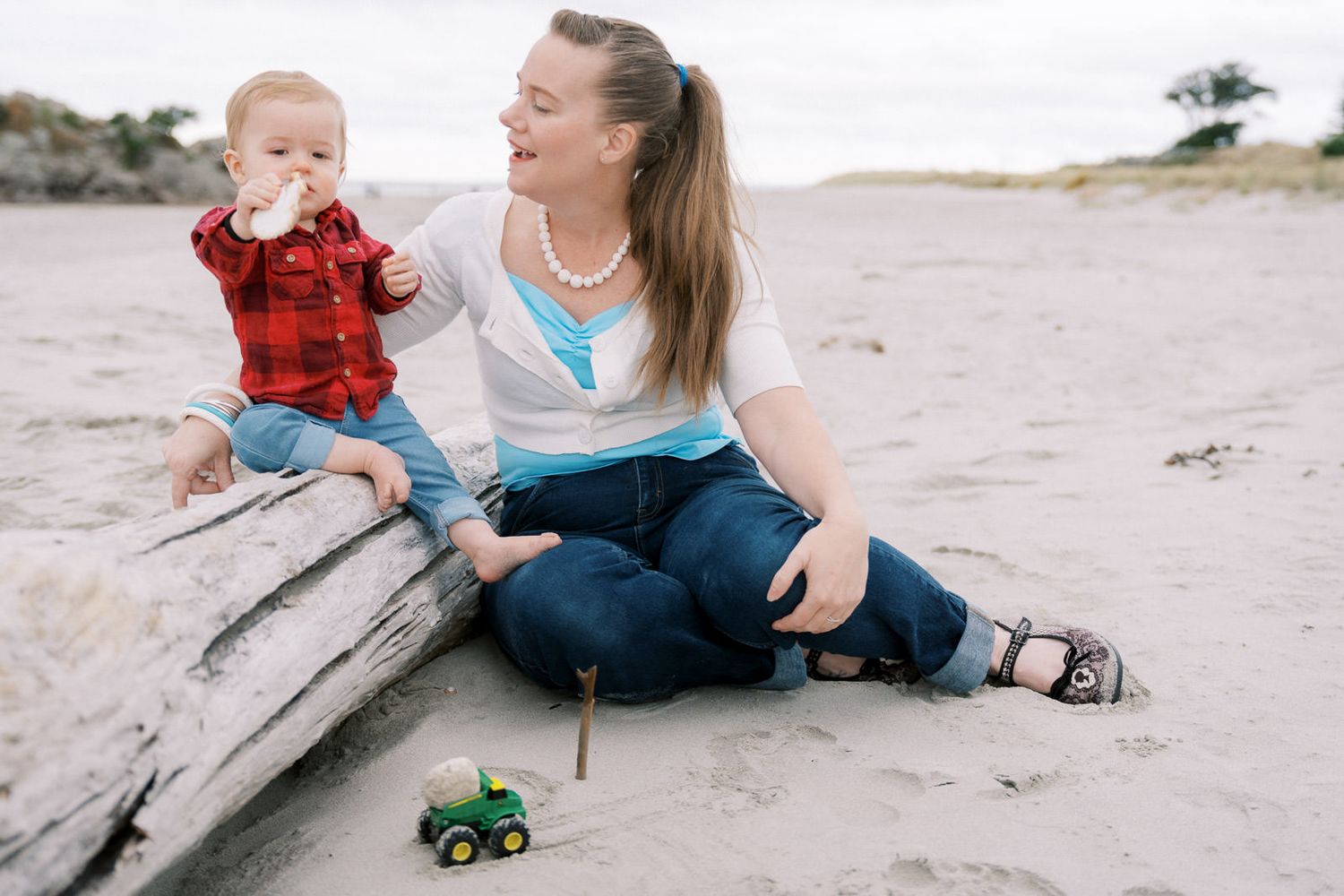 A mother and baby sit on driftwood at the beach, with a toy tractor in the sand nearby.