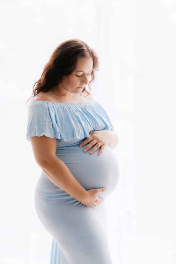 Pregnant woman in light blue off-shoulder dress cradling baby bump, maternity photography studio portrait.