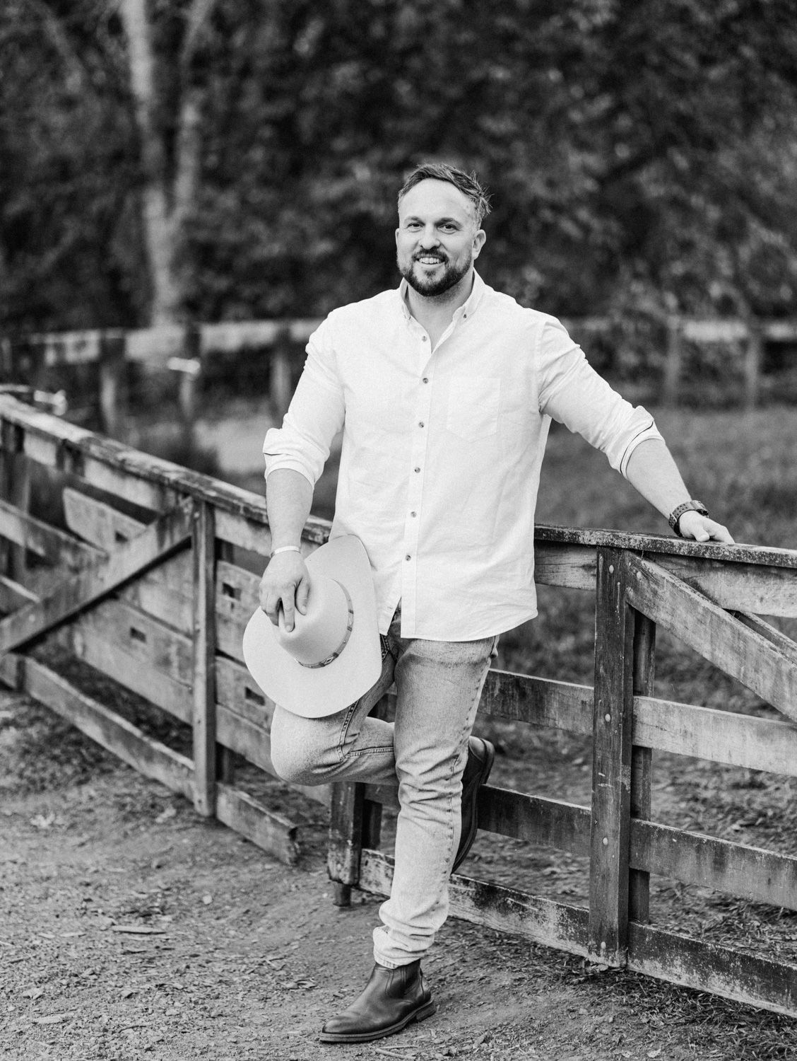 Man in white shirt holding cowboy hat leaning on wooden fence rail in black and white outdoor photo.