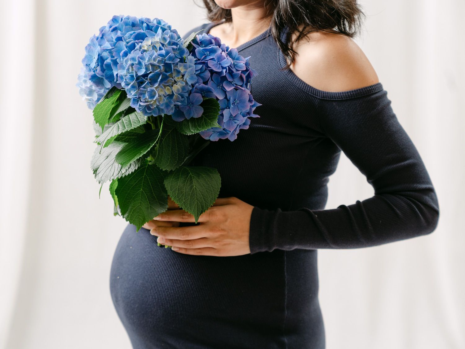 Pregnant woman in navy blue off-shoulder dress holding blue hydrangea flowers against white background.
