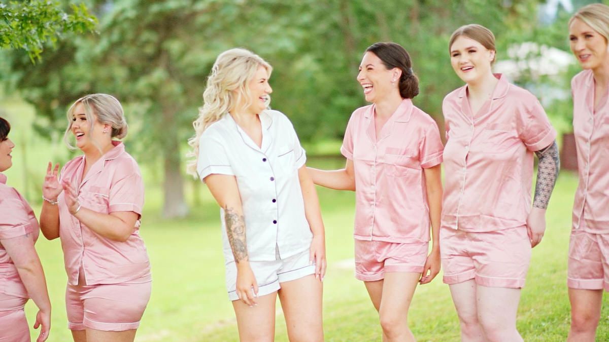 Bride in white pajamas walks with bridesmaids in matching pink satin shorts sets outdoors on a green lawn.
