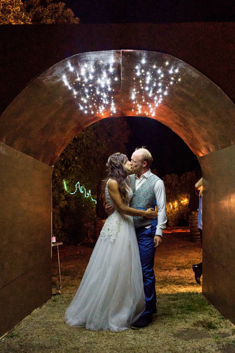 Bride and groom share a romantic kiss under a rustic stone arch with sparkling fireworks at their outdoor wedding reception.