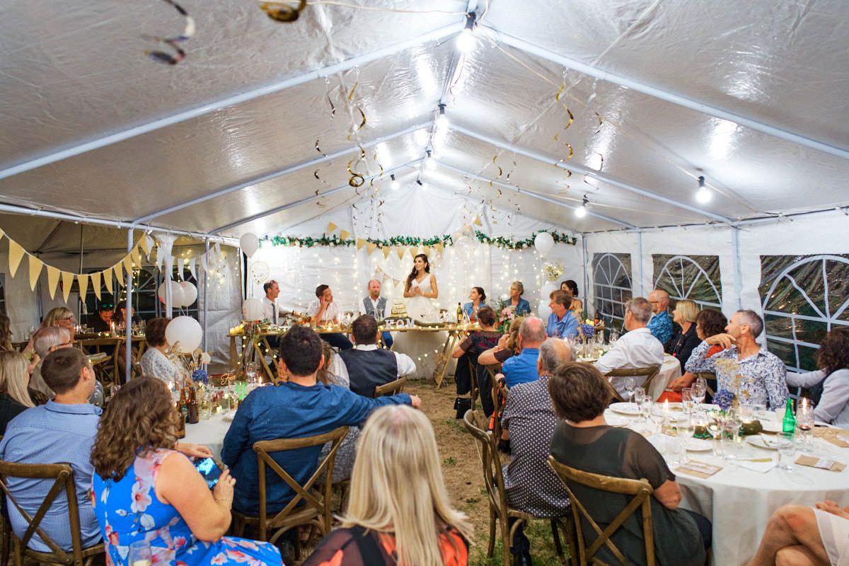 Guests seated at round tables during an elegant outdoor wedding reception under a white tent with decorative lights.