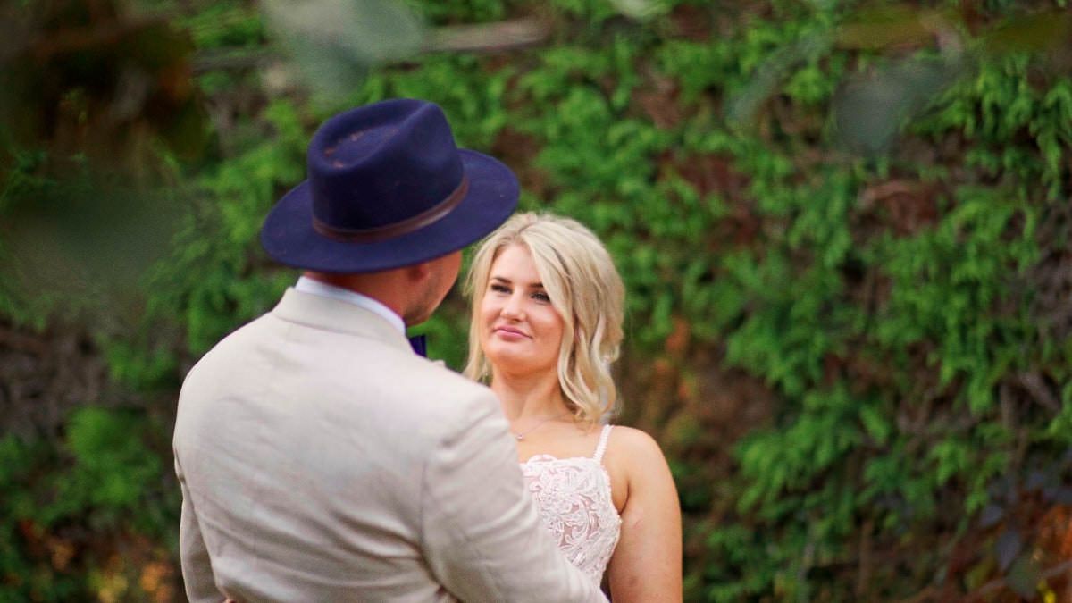 Couple sharing a romantic moment outdoors, bride in lace dress smiles at groom wearing navy hat and gray suit.
